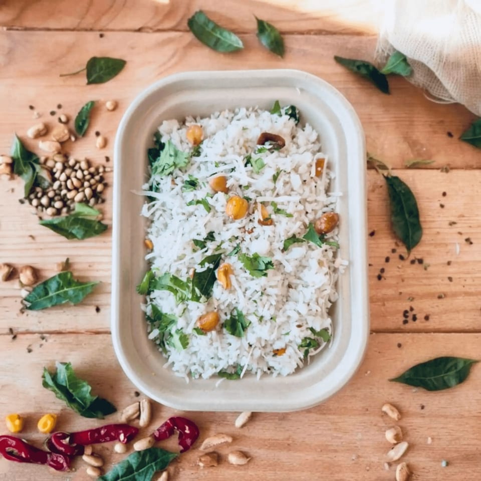 Kolapasi Indian Canteen's coconut rice with peanuts and herbs in a white container on a wooden table.