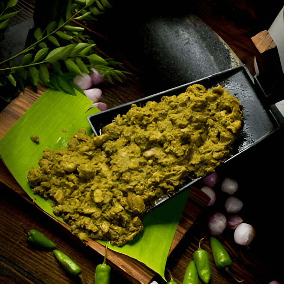 Green Indian curry on a black tray and banana leaf, with chilies, onions, and curry leaves.