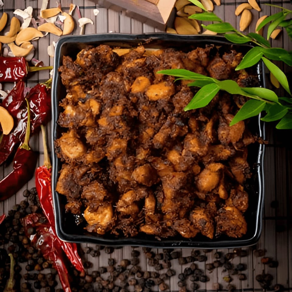 A black dish filled with Kolapasi Indian Canteen's dark, spicy chicken, surrounded by red chilies and peppercorns.