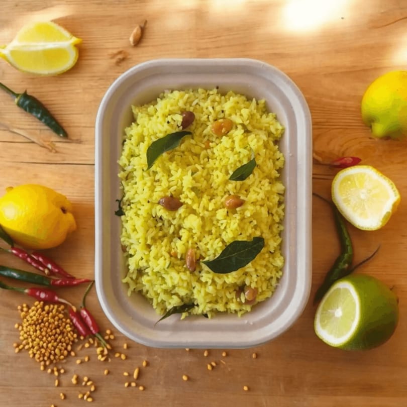 Lemon rice with peanuts and curry leaves in a container, surrounded by lemons, limes, and chilies.